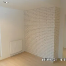 Białe wnętrze po remoncie: ściana z tapetą imitującą cegłę, jasna podłoga, okno z widokiem na zewnątrz i grzejnik. Minimalistyczny design i świeżość.