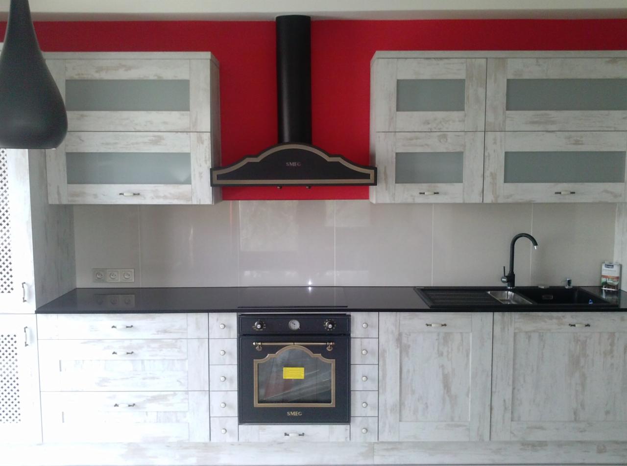 Wykończona kuchnia w stylu rustykalnym z białymi szafkami i czarnym blatem, na tle czerwonej ściany. Widoczny piekarnik i okap marki SMEG.