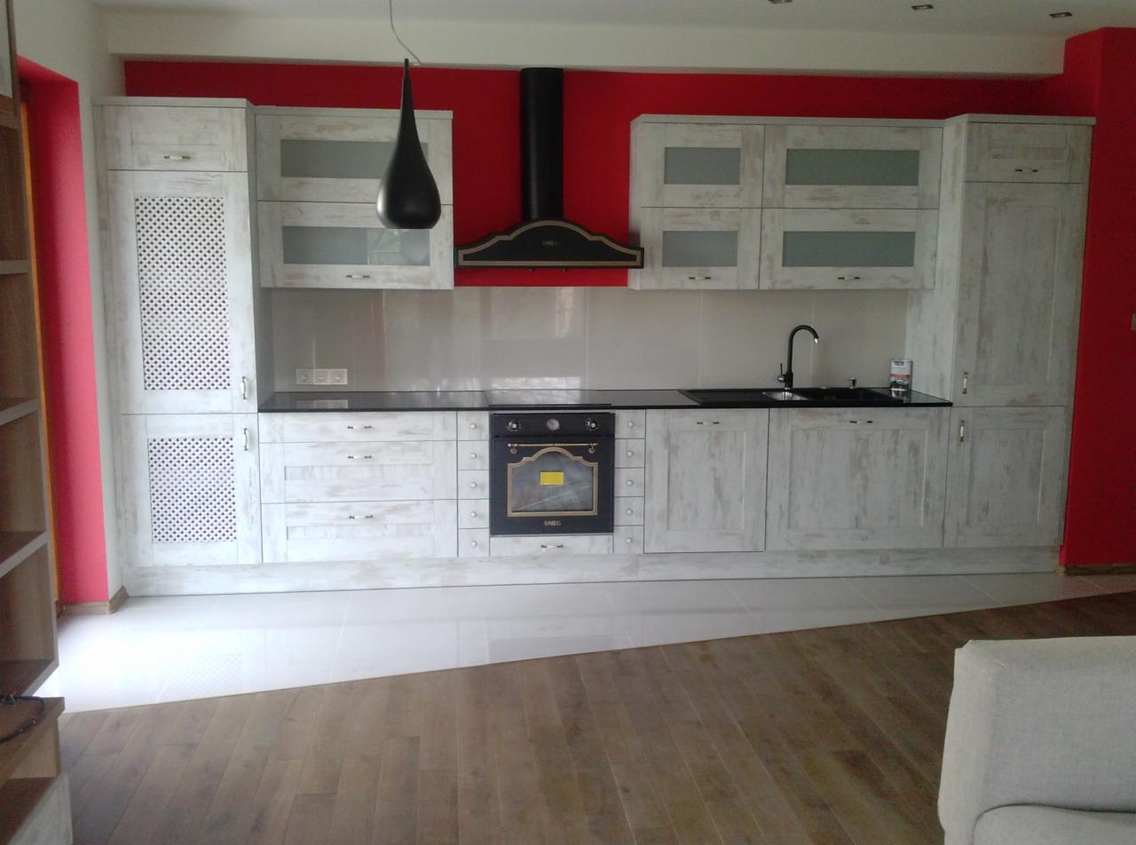 Kuchenna zabudowa w stylu rustykalnym z białymi szafkami i czarnym blatem, na tle czerwonej ściany. Widoczny okap i piekarnik. Podłoga wyłożona płytkami i drewnem.