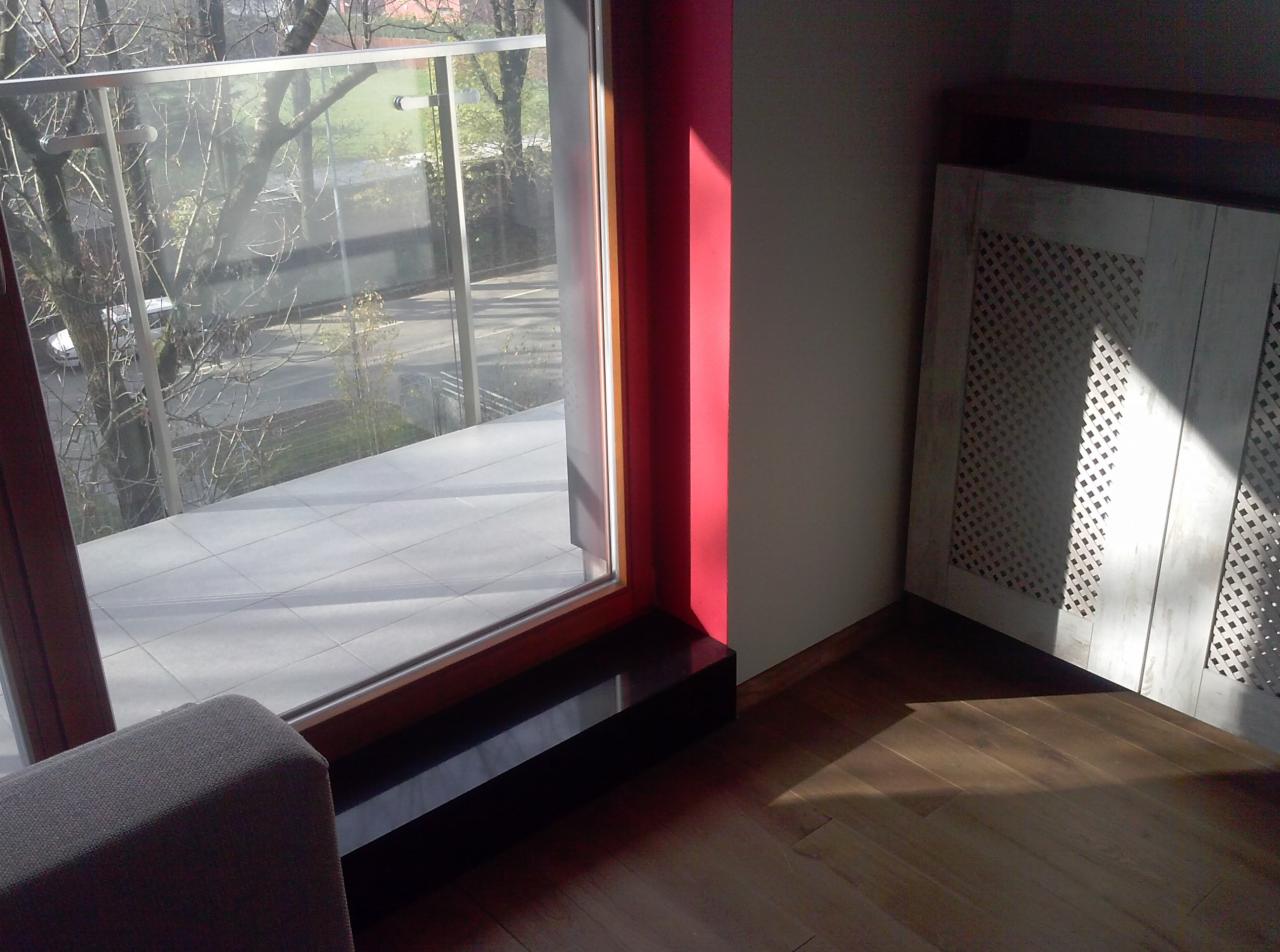 Nowoczesny balkon z szarymi płytkami i szklaną balustradą. Czerwona rama okienna kontrastuje z drewnianą podłogą i dekoracyjnym grzejnikiem w osłonie.