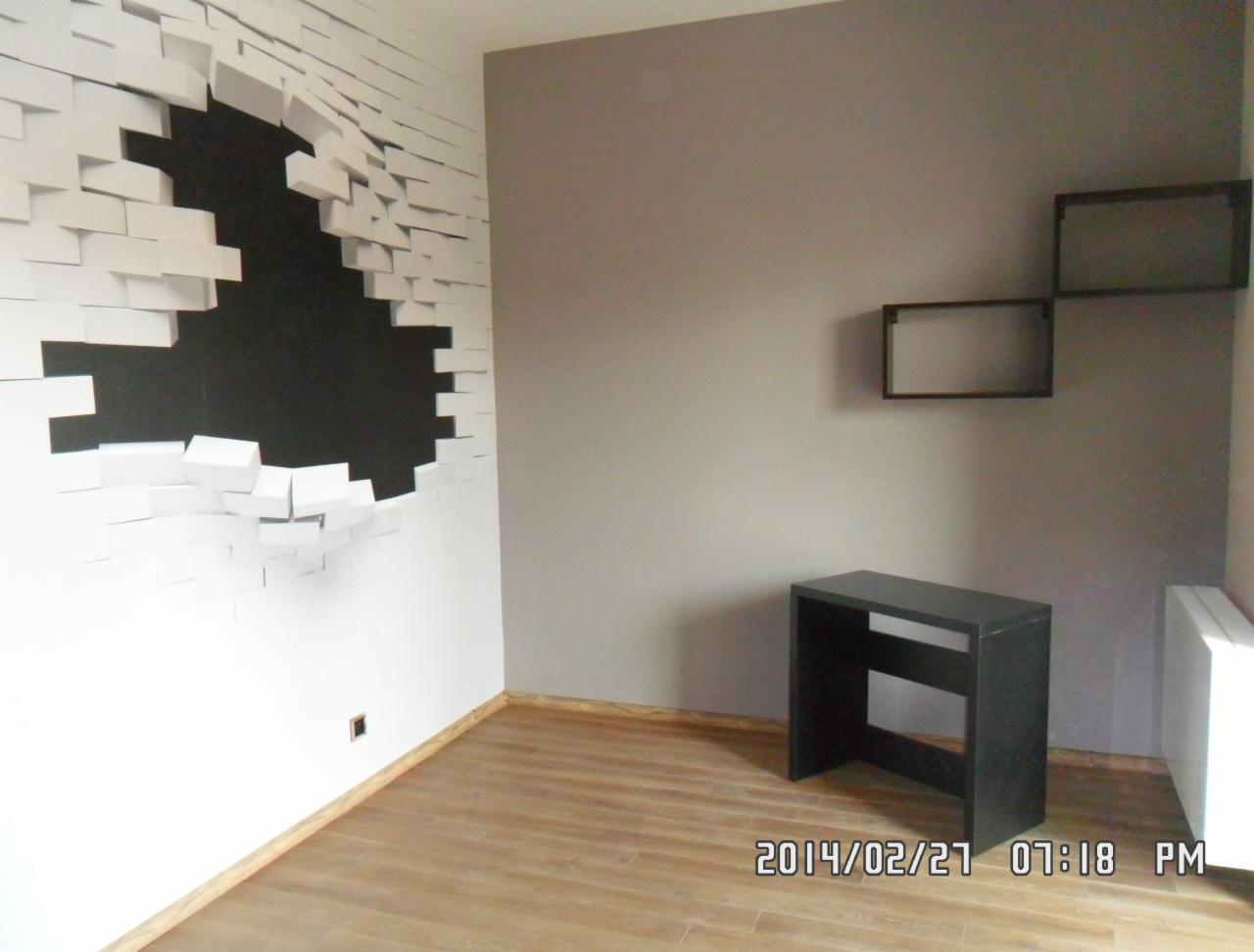 Pokój z dekoracyjną ścianą imitującą rozpadającą się cegłę, drewnianą podłogą i szarą ścianą z minimalistycznymi półkami. Widoczny mały czarny stolik.