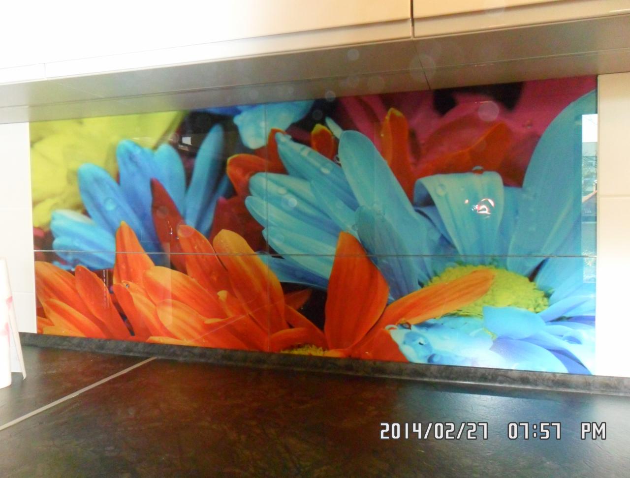 Kolorowa grafika kwiatowa na szklanej powierzchni w kuchni, widoczne łączenia płytek, brązowy blat. Nietypowy wzór na ścianie nad blatem kuchennym.