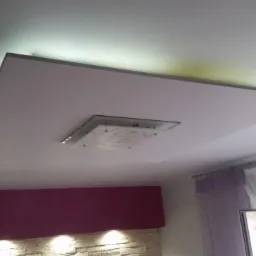 Podwieszany sufit z płyt karton gips z oświetleniem LED i nowoczesną lampą sufitową, widoczna dekoracyjna ściana z kamienia i zasłony w tle.