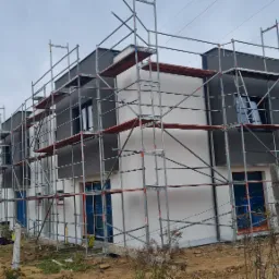 Szereg domów w trakcie budowy, z widocznymi rusztowaniami i częściowo wykonaną elewacją w kolorach szarości i bieli, okna zabezpieczone folią.