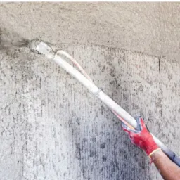 Aplikacja mokrego tynku na chropowatą ścianę za pomocą pistoletu natryskowego, widoczne rękawice ochronne i przewody doprowadzające materiał.