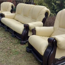 Komplet wypoczynkowy: sofa i dwa fotele z jasnej skóry ekologicznej i ciemnego, lakierowanego drewna, ustawiony na trawie z liśćmi.