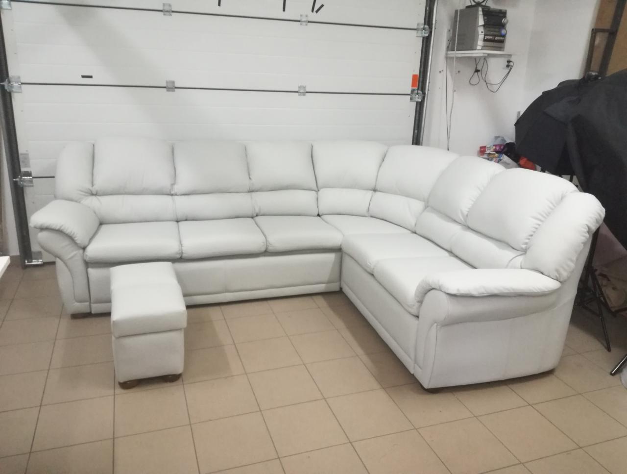 Jasny, narożny zestaw wypoczynkowy z białej skóry, uzupełniony o puf, sfotografowany w garażu z widocznymi drzwiami segmentowymi i sprzętem audio na półce w tle.