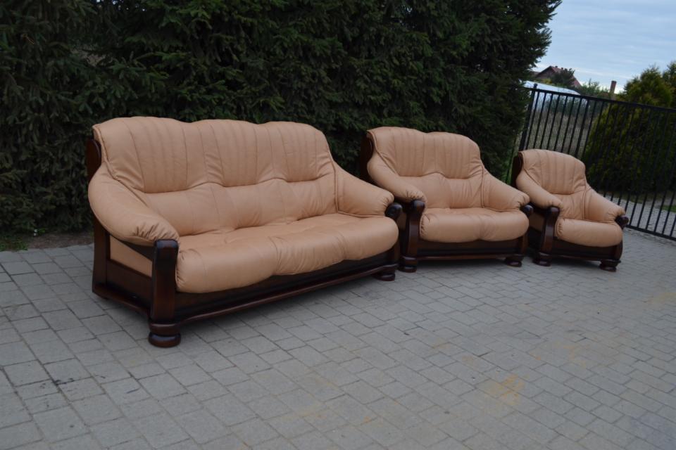 Zestaw wypoczynkowy: sofa trzyosobowa, fotel i mały fotel, w beżowej skórzanej tapicerce i ciemnobrązowych drewnianych ramach, ustawiony na brukowanym podwórku przed zielonymi tujami i czarnym...