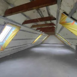 Poddasze ocieplone pianką PUR, widoczne okna dachowe zabezpieczone folią, drewniane elementy konstrukcyjne, szara podłoga.