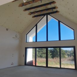 CELS Mariusz Butkiewicz - Wnętrze domu w stanie surowym otwartym, widoczne ocieplone poddasze z odsłoniętą konstrukcją drewnianą, duże okna z widokiem na zewnątrz, instalacje elektryczne wyprowadzone ze ścian.