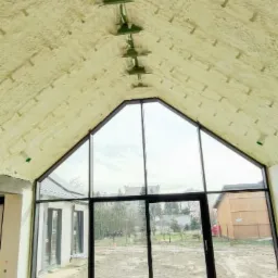 Poddasze domu z widoczną warstwą ocieplenia pianką PUR, przymocowaną plastikowymi kołkami, duże okno z czarnymi ramami z widokiem na ogród.