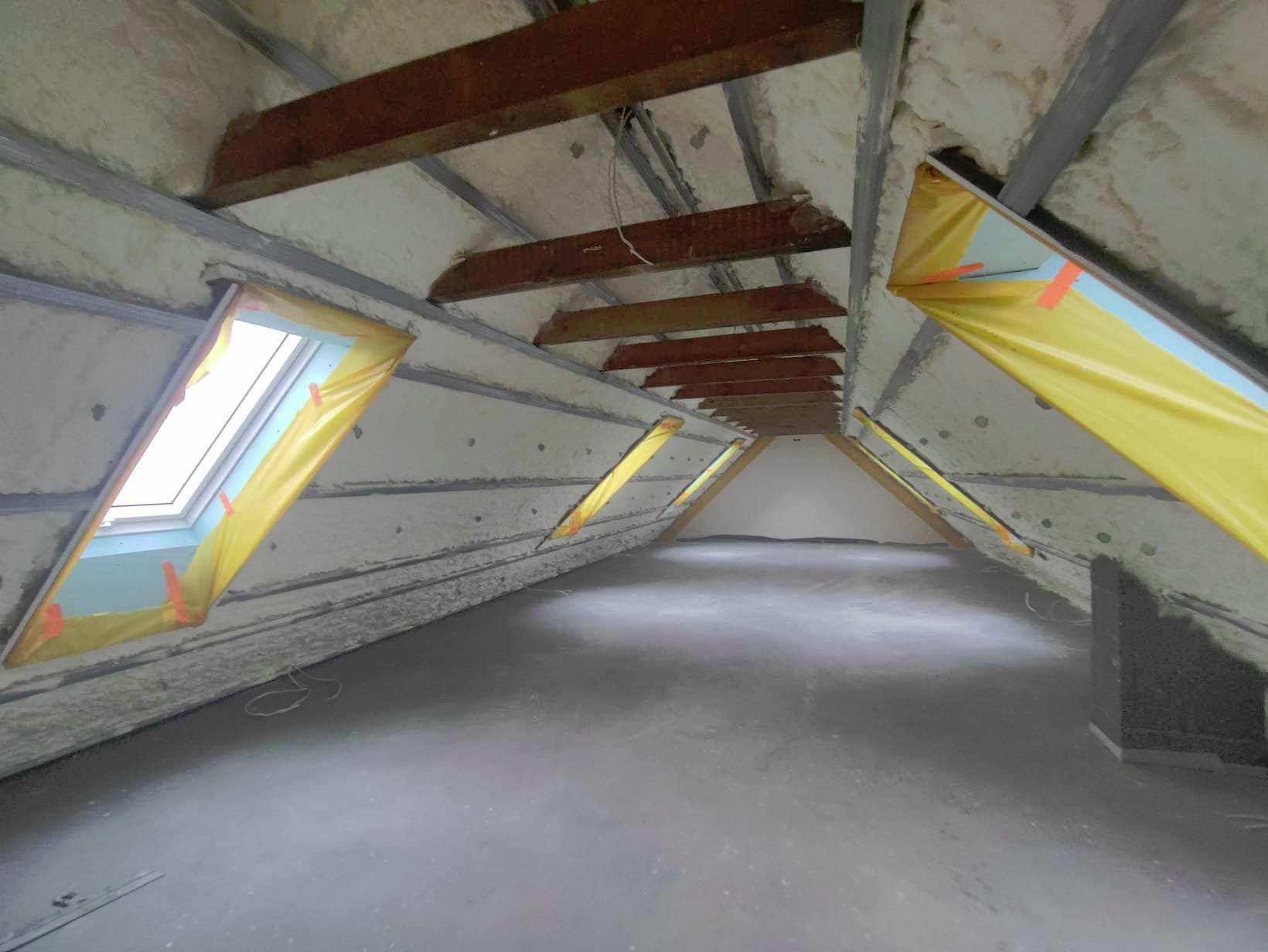 Poddasze ocieplone pianką PUR, widoczne okna dachowe zabezpieczone folią, drewniane elementy konstrukcyjne, szara podłoga.