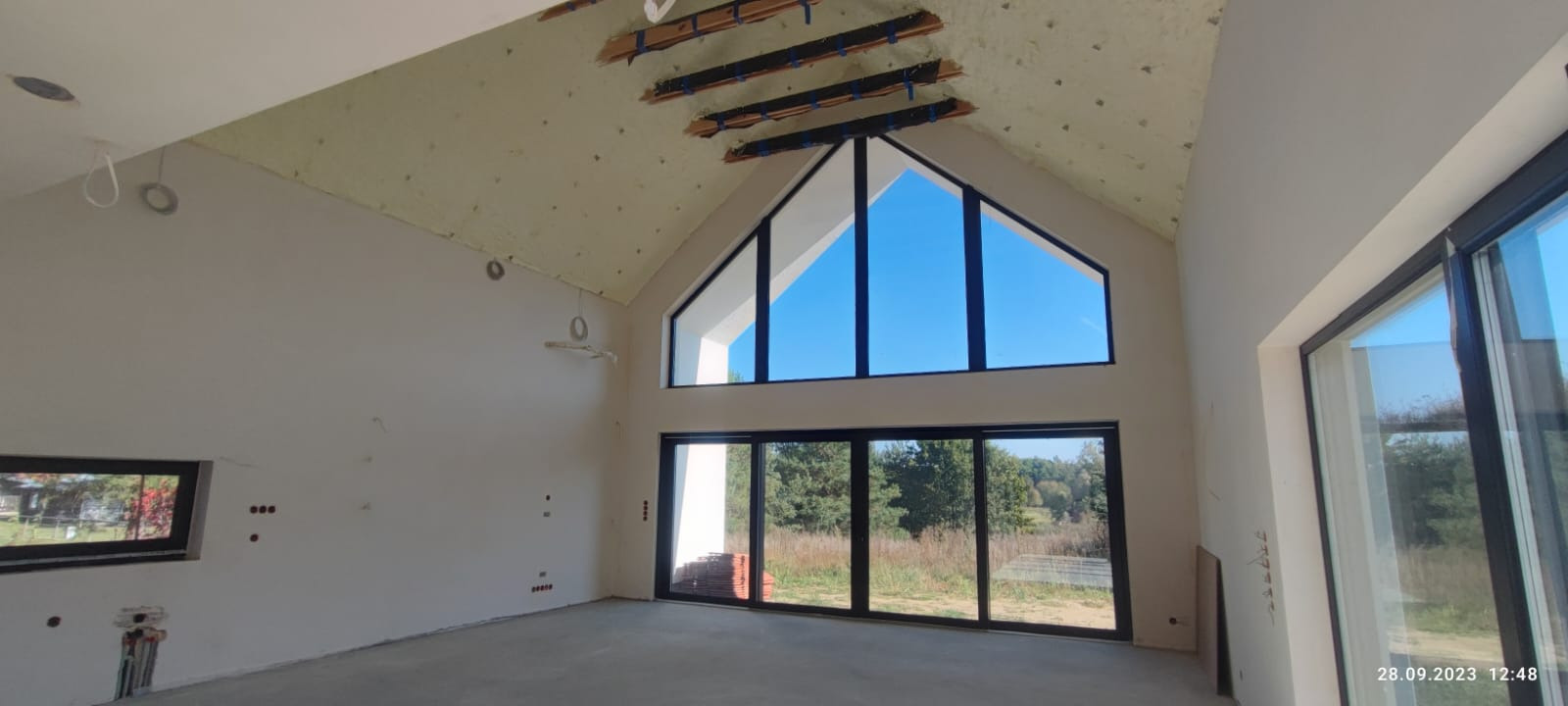 Wnętrze domu w stanie surowym otwartym, widoczne ocieplone poddasze z odsłoniętą konstrukcją drewnianą, duże okna z widokiem na zewnątrz, instalacje elektryczne wyprowadzone ze ścian.