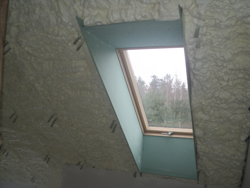 Poddasze ocieplone pianką PUR wokół okna dachowego, widoczne metalowe mocowania, drzewa w tle za oknem.