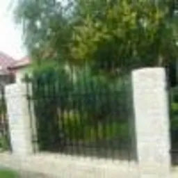 Ciemne, metalowe przęsło ogrodzeniowe z pionowymi szczeblami, osadzone pomiędzy dwoma jasnymi, murowanymi słupkami. W tle widoczne drzewa i fragment dachu budynku.