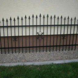 Fragment dekoracyjnego, czarnego ogrodzenia metalowego z motywami roślinnymi, ustawionego przy beżowej ścianie budynku i szarym żwirze.