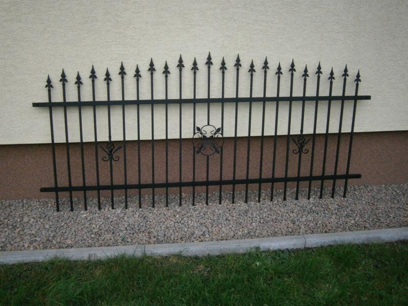 Fragment dekoracyjnego, czarnego ogrodzenia metalowego z motywami roślinnymi, ustawionego przy beżowej ścianie budynku i szarym żwirze.