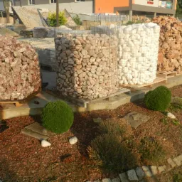 Palety z kamieniami ogrodowymi o różnych kolorach i fakturach, umieszczone na terenie szkółki roślin. Dekoracyjne krzewy i wykończenie z kory.