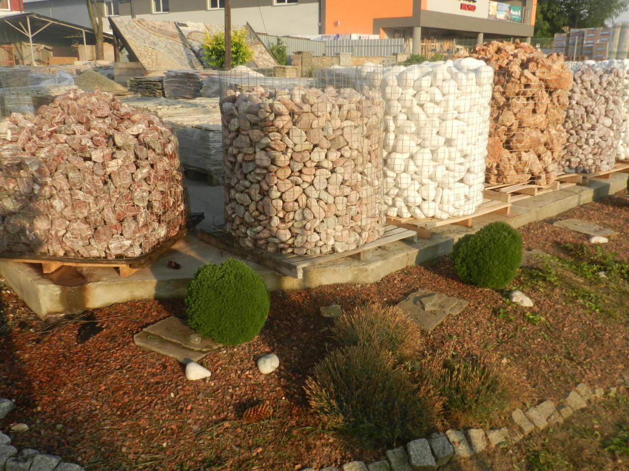 Palety z kamieniami ogrodowymi o różnych kolorach i fakturach, umieszczone na terenie szkółki roślin. Dekoracyjne krzewy i wykończenie z kory.