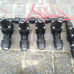Rozdzielacz do systemów nawadniania z pięcioma elektrozaworami połączonymi przewodami elektrycznymi, ułożony na szarej kostce brukowej.