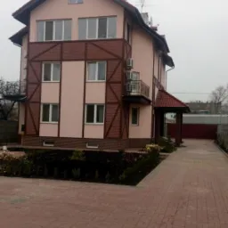 Dwupiętrowy dom z różową elewacją i ceglanymi wstawkami, widziany z perspektywy chodnika z czerwonej kostki. Widoczne balkony i wejście zadaszone czerwoną dachówką.