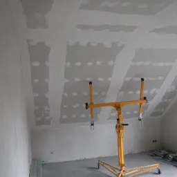 Montaż płyt karton-gips na poddaszu ze skosami. Żółty podnośnik do płyt, widoczne łączenia płyt wypełnione gipsem, przygotowane do szlifowania.