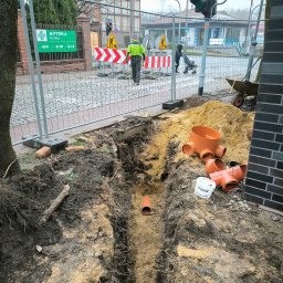 WIRMAX ROMAN LIWA, JAROSŁAW WRÓBEL SPÓŁKA CYWILNA - Wykop w ziemi z widocznymi rurami kanalizacyjnymi z tworzywa sztucznego, zabezpieczony siatką, w tle ulica z sygnalizacją świetlną i przechodniami.