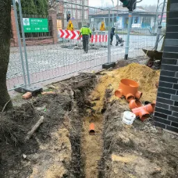 Wykop w ziemi z widocznymi rurami kanalizacyjnymi z tworzywa sztucznego, zabezpieczony siatką, w tle ulica z sygnalizacją świetlną i przechodniami.