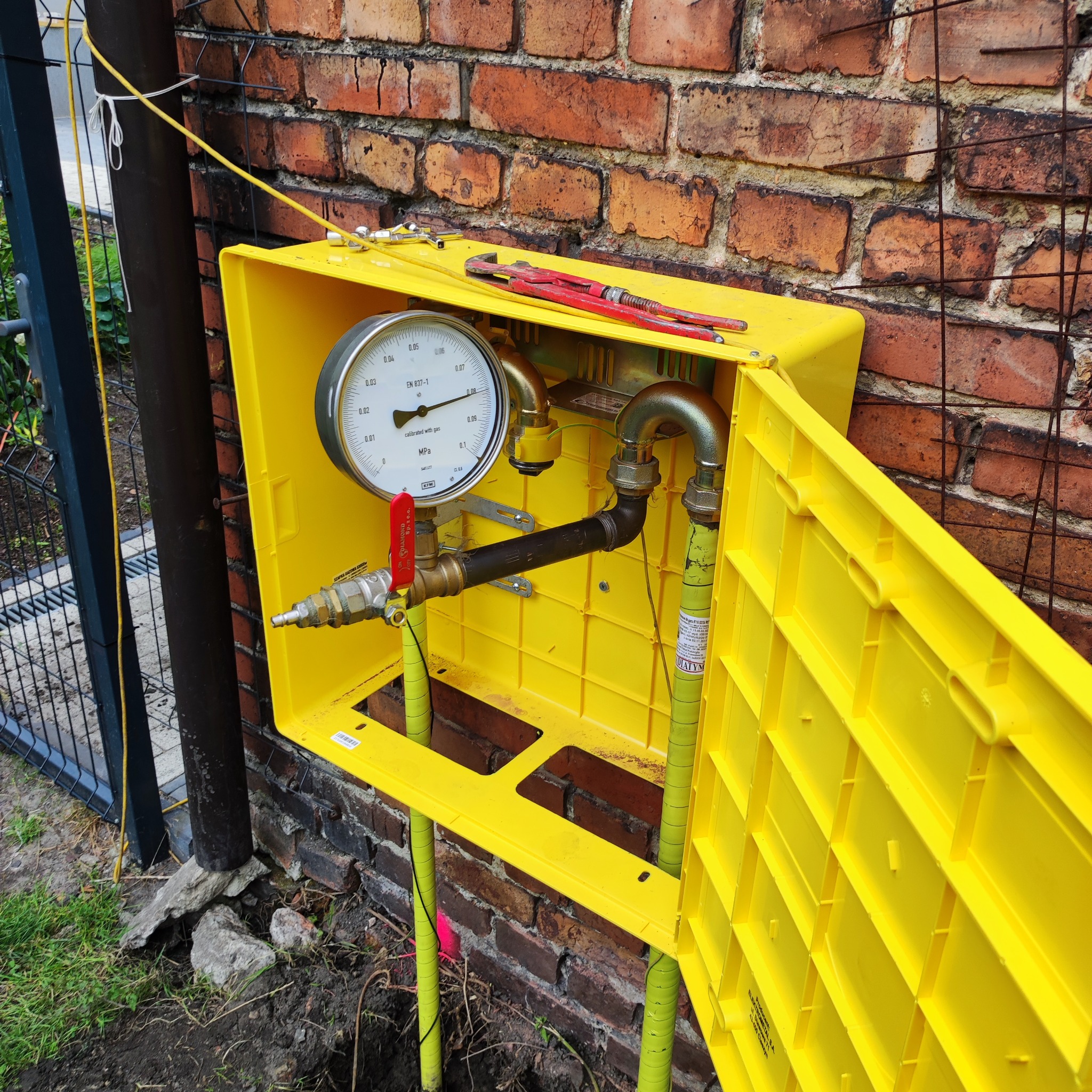 Żółta skrzynka gazowa z manometrem i zaworem, zamontowana na zewnątrz budynku z cegły, widoczne rury gazowe i klucz nastawny na górze skrzynki.