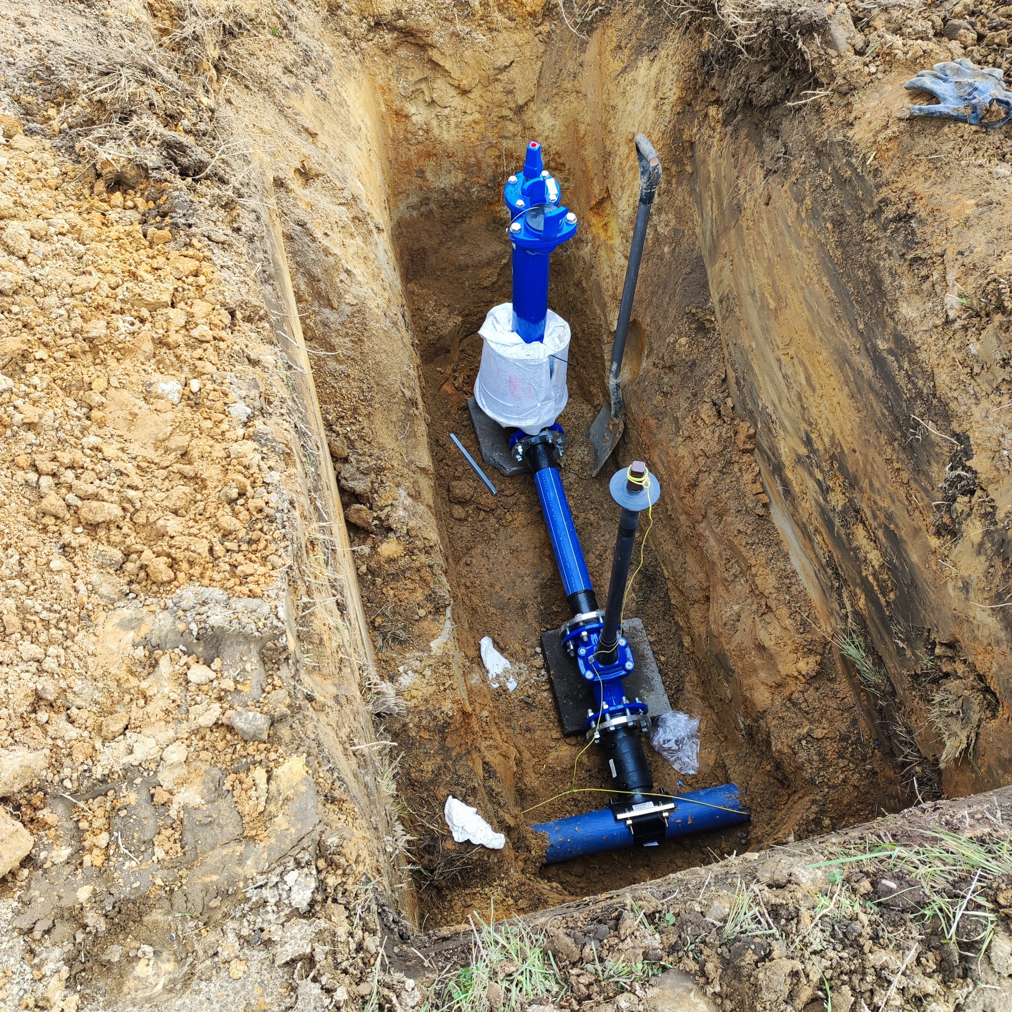 Głęboki wykop z ułożonym niebieskim rurociągiem, częściowo owiniętym folią ochronną, widoczna łopata i narzędzia, ziemia o zróżnicowanej strukturze.