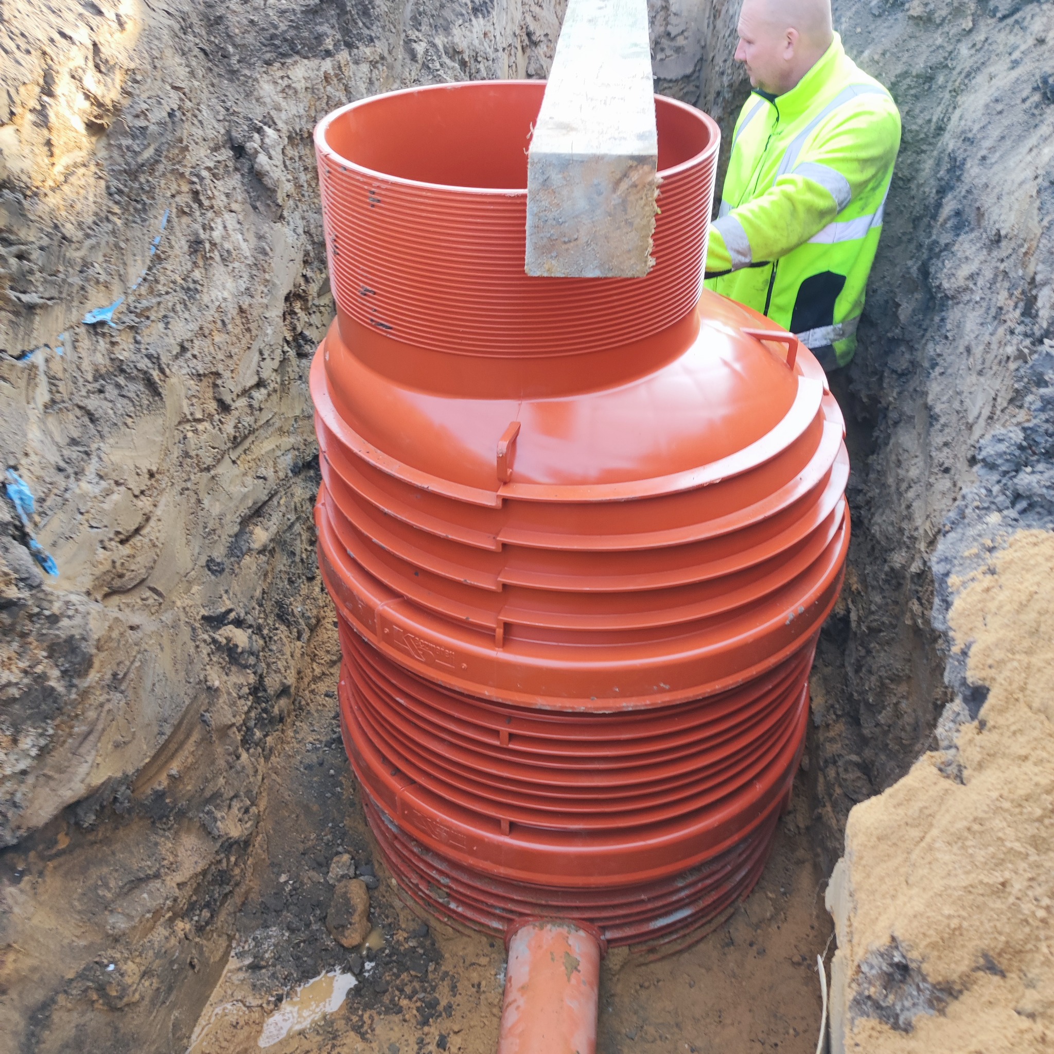Pomarańczowy zbiornik z tworzywa sztucznego, z widocznym przyłączem rury, umieszczony w wykopie ziemnym. Pracownik w odblaskowej kurtce nadzoruje montaż.