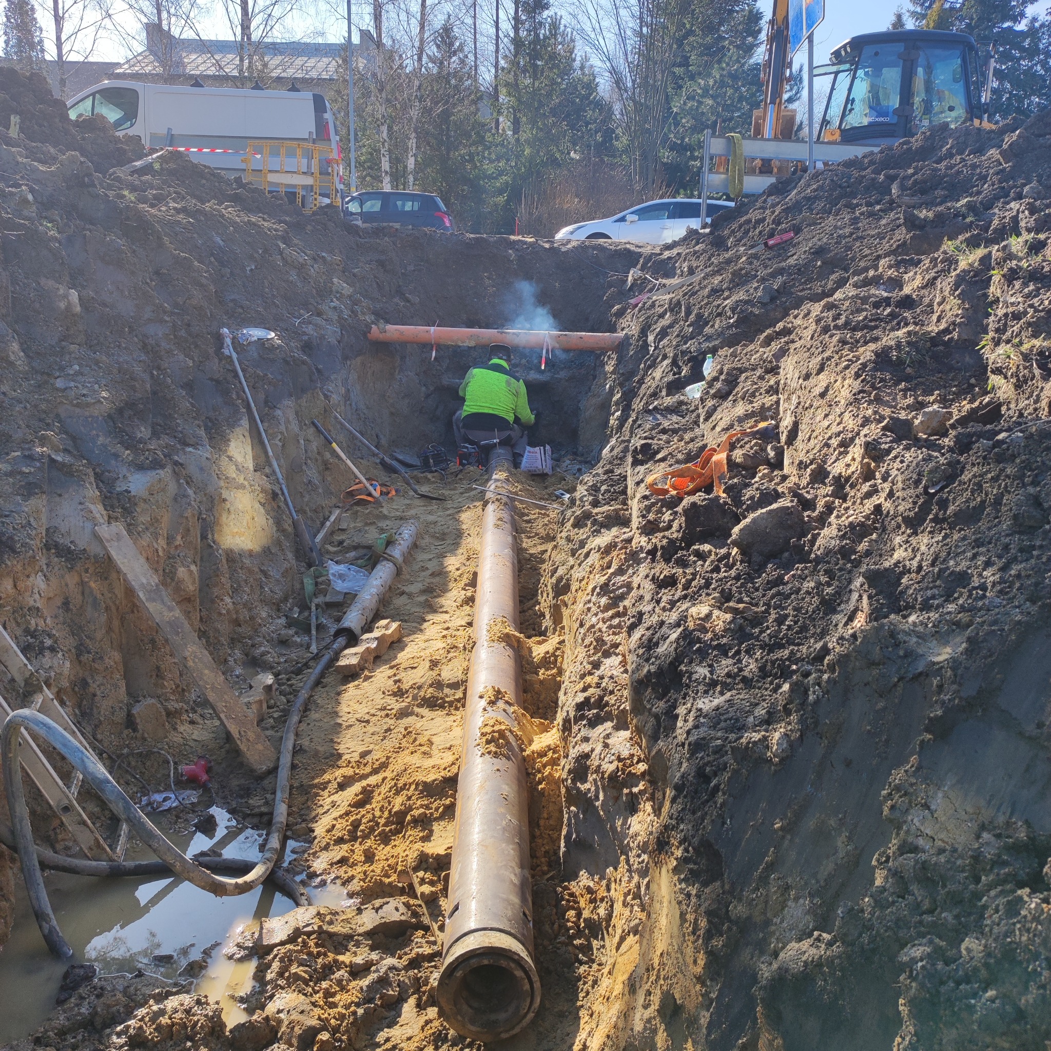Głęboki wykop z widoczną rurą kanalizacyjną, pracownik w zielonej kurtce łączy rury, w tle sprzęt budowlany i samochody.