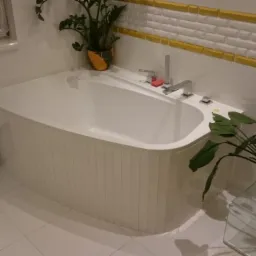 Nowoczesna łazienka z białą wanną obudowaną płytkami, dekoracyjną ścianą w odcieniach bieli i złota oraz roślinami doniczkowymi. Uwagę zwraca waga łazienkowa.