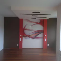 Wnętrze pokoju z drewnianą podłogą, brązową ścianą i dekoracyjną ścianą z czerwonymi elementami. Widoczny fragment okna i drzwi.