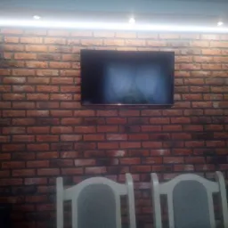 Ściana z cegły z zamontowanym telewizorem i oświetleniem LED podsufitowym, widoczne dwa krzesła z jasnego drewna.