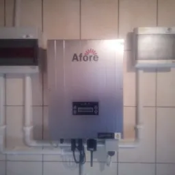 Szary falownik z logo Afore, zamontowany na kafelkowanej ścianie, z podłączonymi kablami i rurami, obok dwie skrzynki elektryczne.