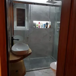 Nowoczesna łazienka z kabiną prysznicową typu walk-in, umywalką nablatową na drewnianym blacie i szarą glazurą imitującą marmur, oświetlona punktowo.