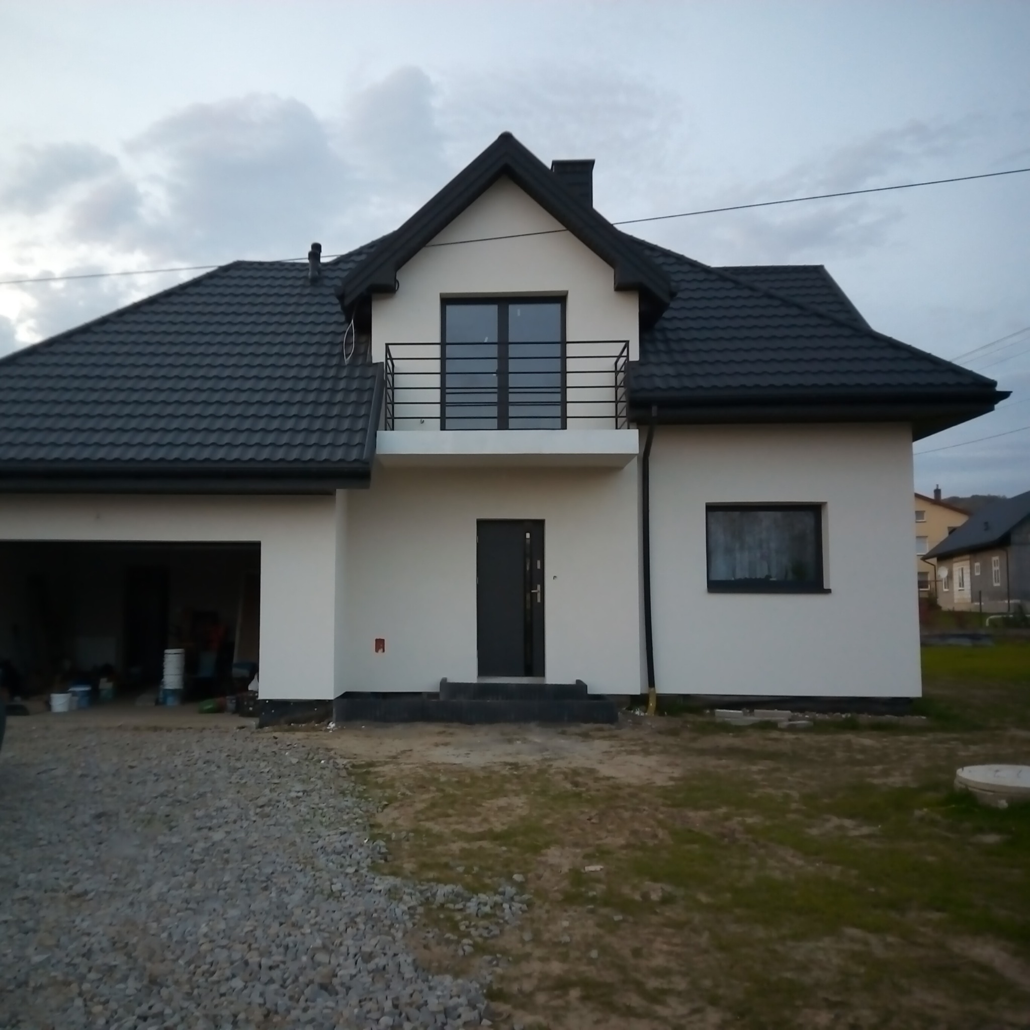Nowoczesny dom jednorodzinny z czarnym dachem i białymi ścianami, widok z zewnątrz na podjazd z kamieniami, z balkonem i drzwiami wejściowymi.