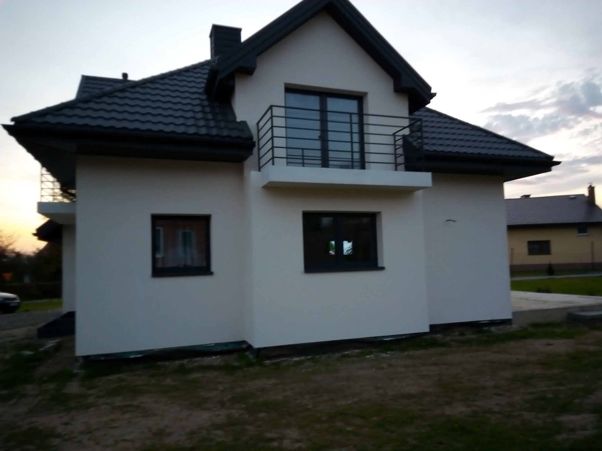 Nowoczesny dom jednorodzinny z białymi ścianami, ciemną dachówką i balkonem z czarną balustradą, widoczny z boku na tle zachodzącego słońca.