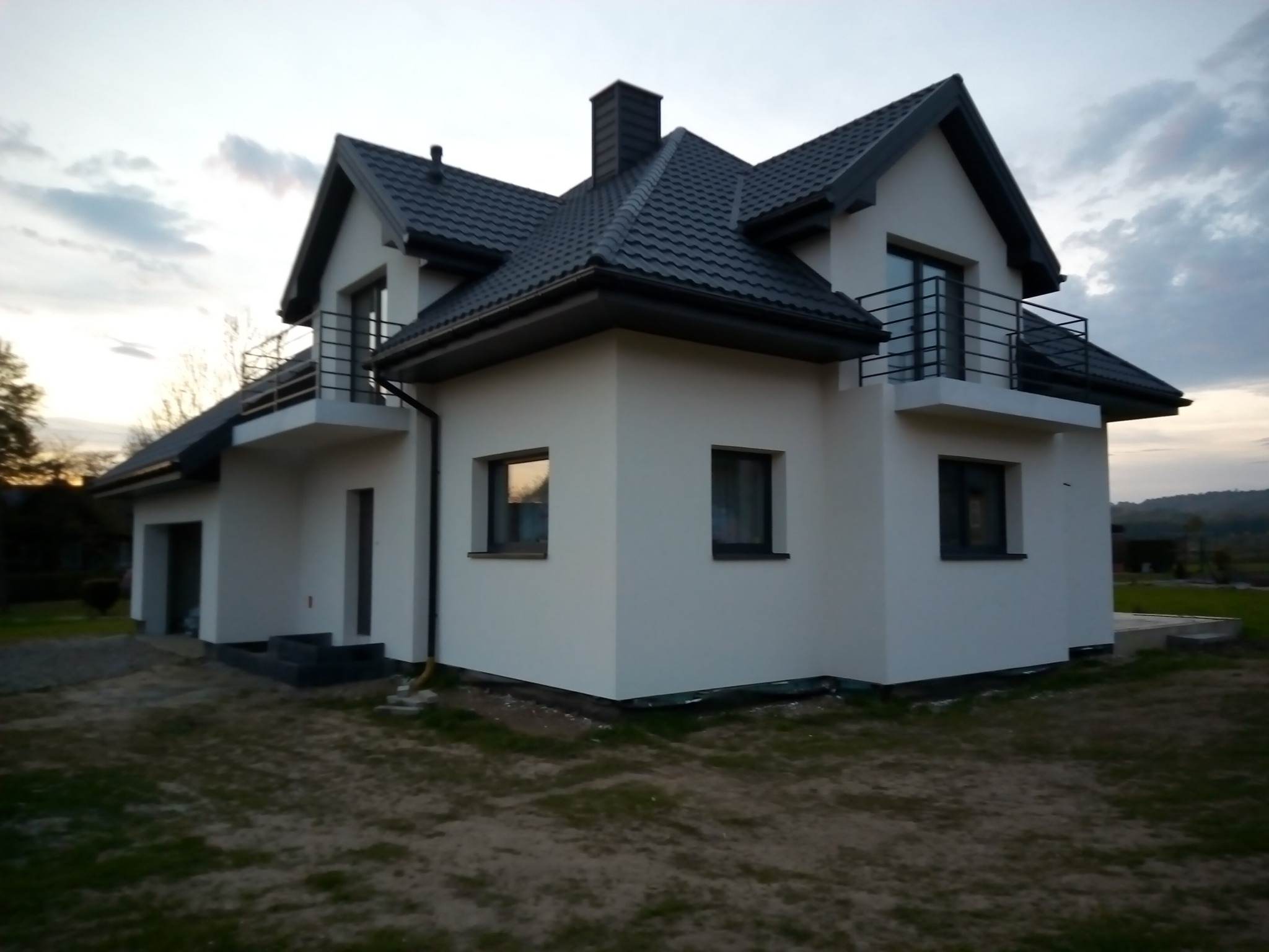 Nowoczesny dom jednorodzinny z dwoma balkonami, wykończony w bieli i szarości, widoczny o zmierzchu na tle pagórkowatego krajobrazu.