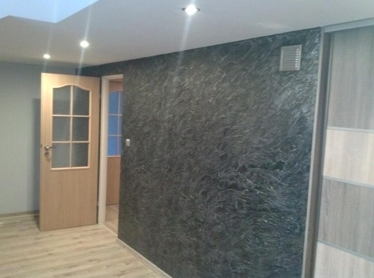 Ściana z efektem dekoracyjnym w odcieniach szarości, widoczny fragment podłogi, drzwi wejściowe i przesuwne drzwi szafy. Oświetlenie sufitowe.