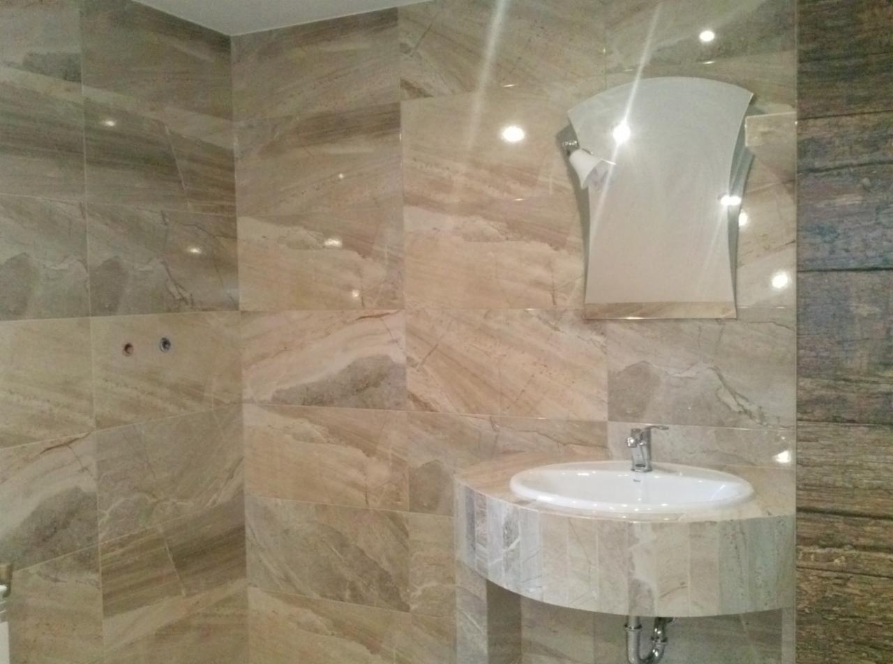 Wykończona łazienka z umywalką wpuszczoną w blat z płytek imitujących marmur, lustro o nietypowym kształcie i płytki ścienne w podobnym stylu.