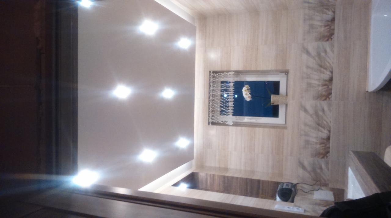 Nowoczesna łazienka z beżowymi płytkami imitującymi drewno, oświetleniem punktowym na suficie i oknem z dekoracyjną zasłoną. Widoczny fragment wanny.