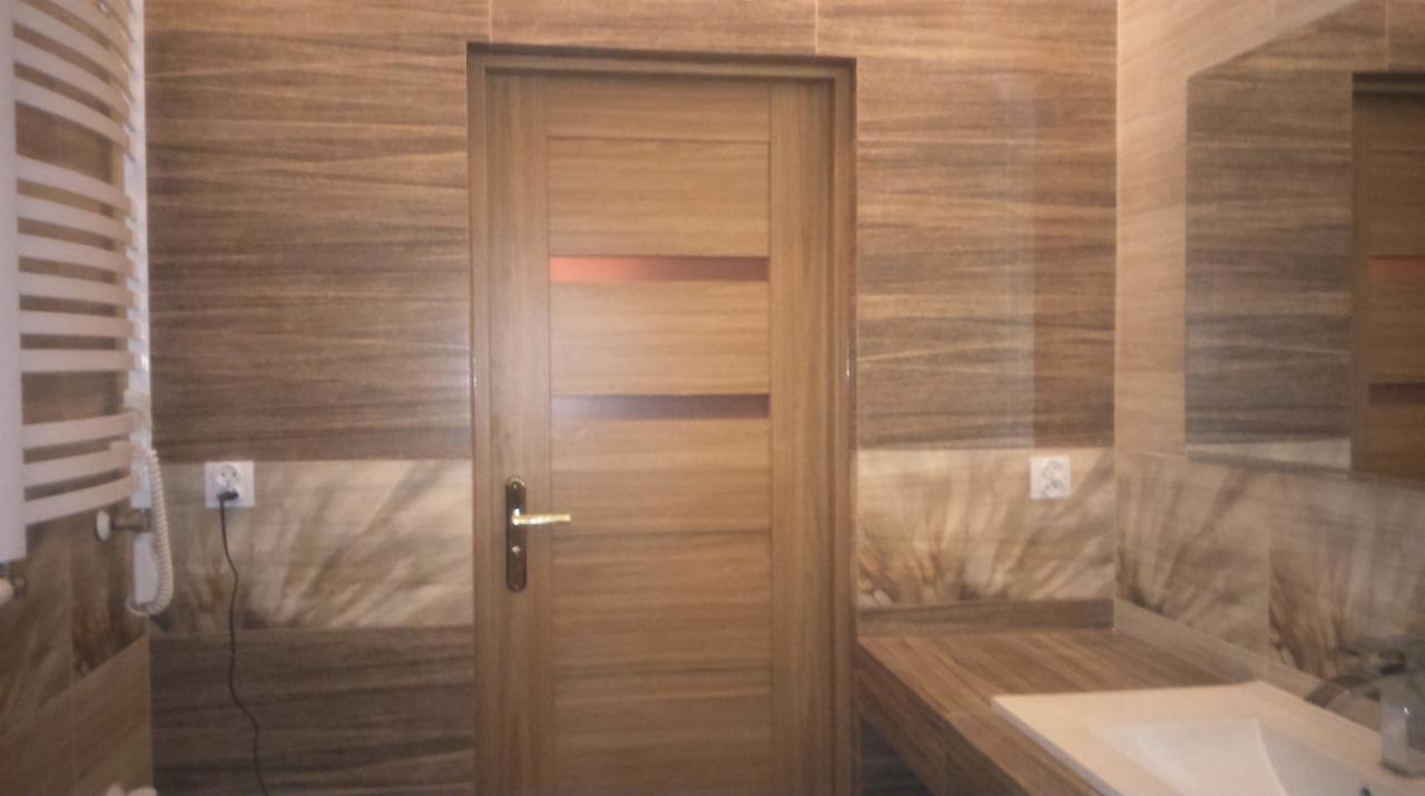 Wykończona łazienka z drewnopodobnymi płytkami, zabudową umywalki i drzwiami z poziomymi przeszkleniami. Widoczny grzejnik drabinkowy i lustro.