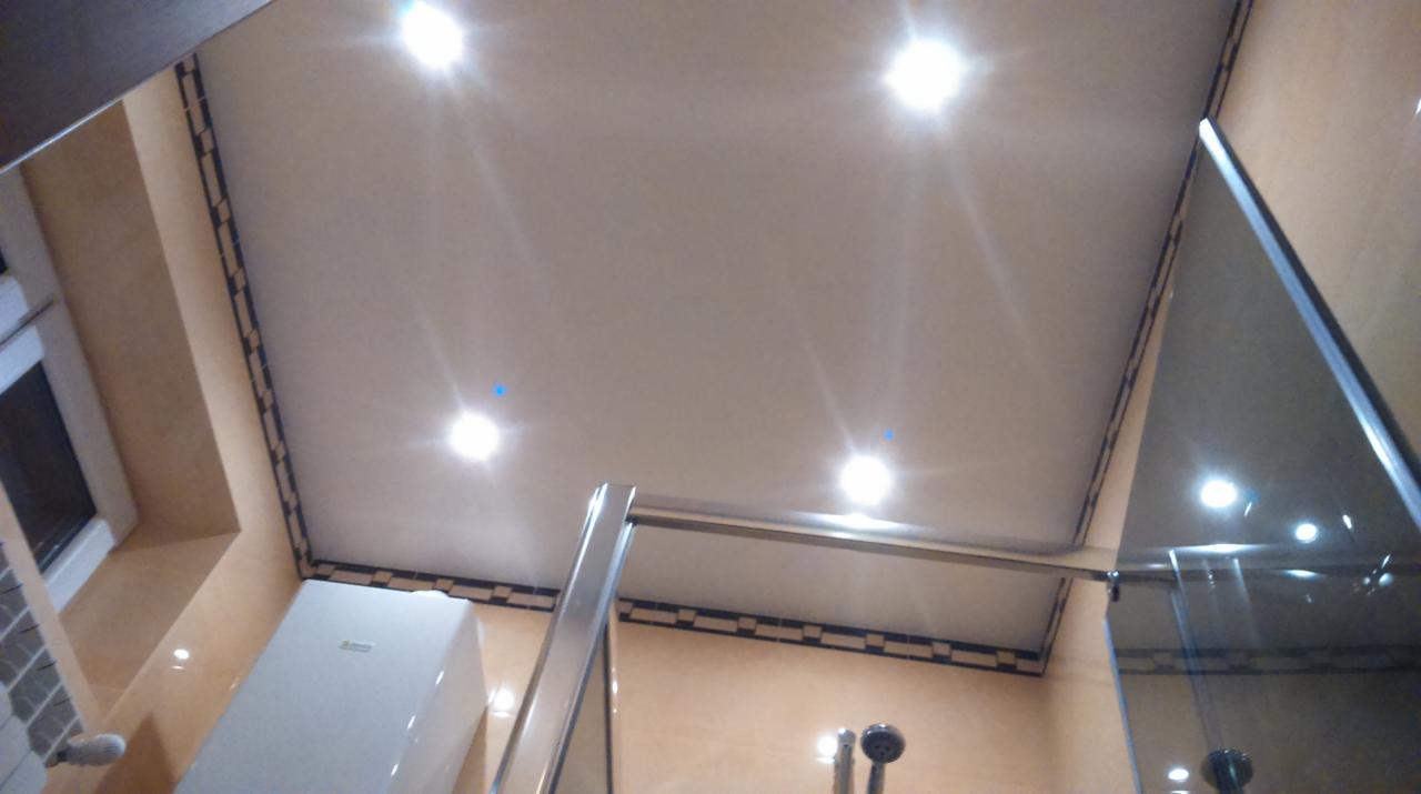 Jasne oświetlenie w łazience z kremowymi płytkami, kabiną prysznicową i podwieszanym sufitem z dekoracyjną obwódką. Widok z dołu.