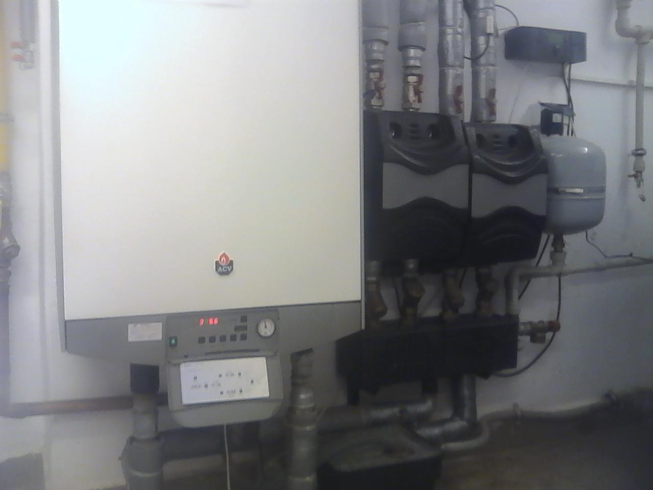 Kocioł grzewczy ACV zamontowany na ścianie, widoczne rury, zawory i inne elementy instalacji grzewczej w piwnicy lub kotłowni. Na kotle widoczny wyświetlacz i panel sterowania.