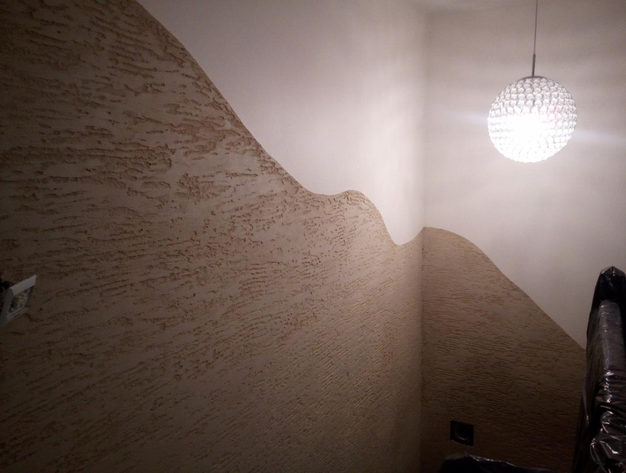 Dekoracyjna ściana z fakturą przypominającą piasek, z falistym przejściem do gładkiej, białej powierzchni, oświetlona przez kryształową lampę wiszącą.