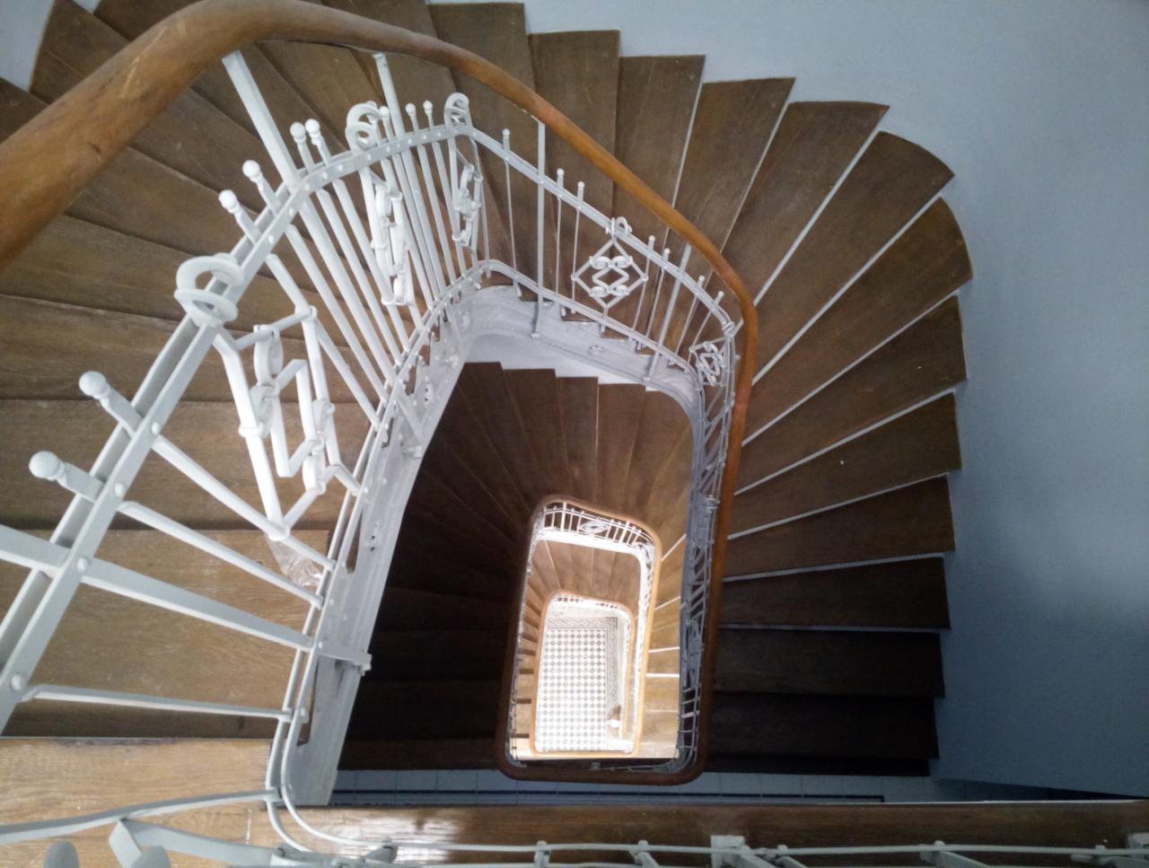 Klatka schodowa z drewnianymi schodami i białą, metalową balustradą, widok z góry w dół, z widocznym wzorem podłogi na najniższym poziomie.