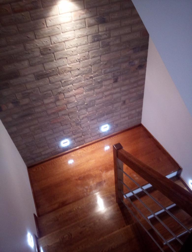 Wykończone schody z drewnianymi stopniami i metalową balustradą, ściana wyłożona cegłą dekoracyjną z wbudowanym oświetleniem punktowym.