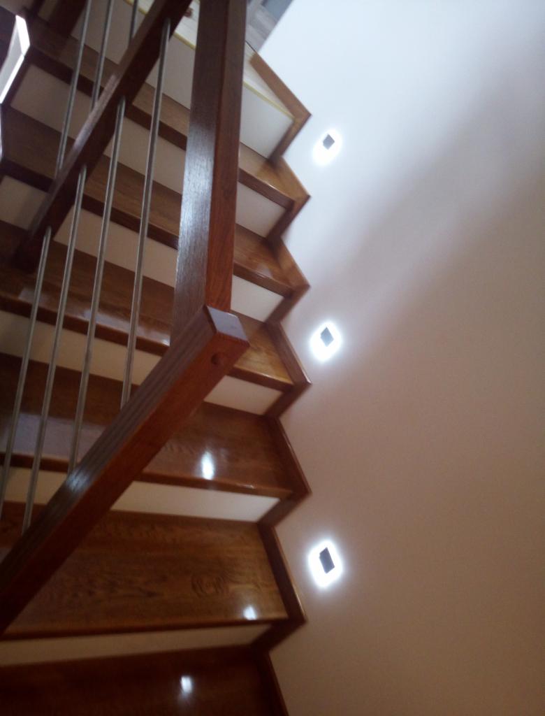 Drewniane schody z metalową balustradą, oświetlone punktowymi lampkami w ścianie, widok z dołu.